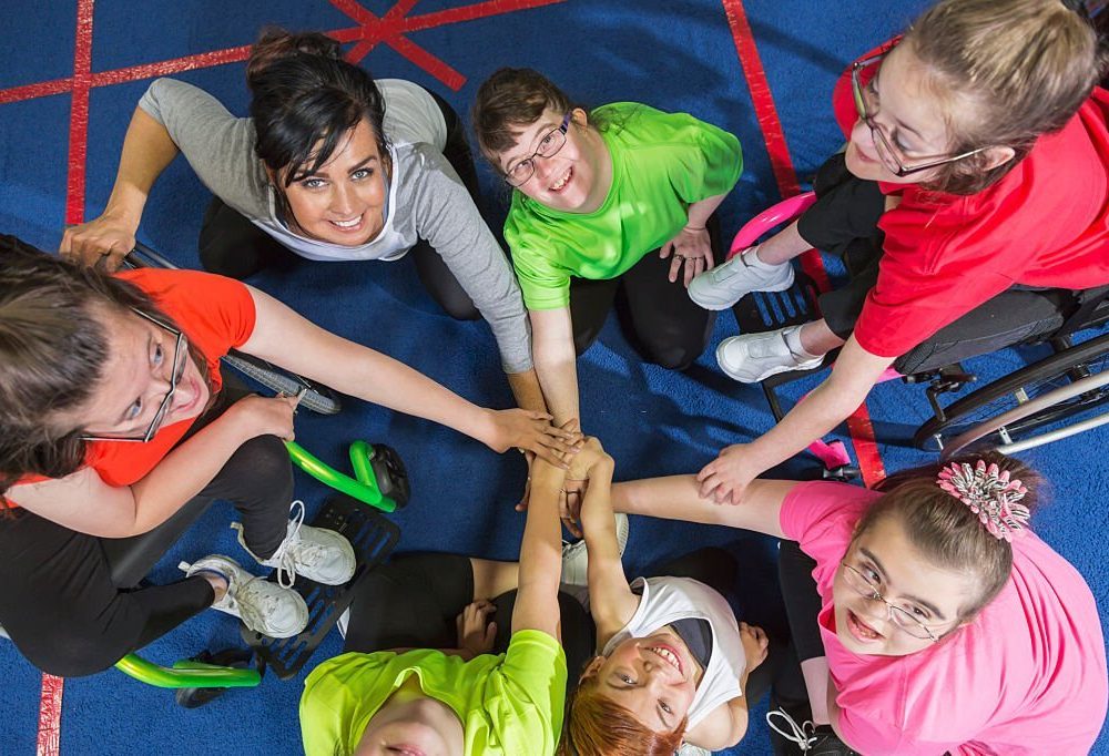 Overhead view of a group of special needs teenagers and young women with instructor, showing team spirit. They are sitting in a circle on a gym floor, hands in the center. Several of the girls have downs syndrome and two are in wheelchairs.
