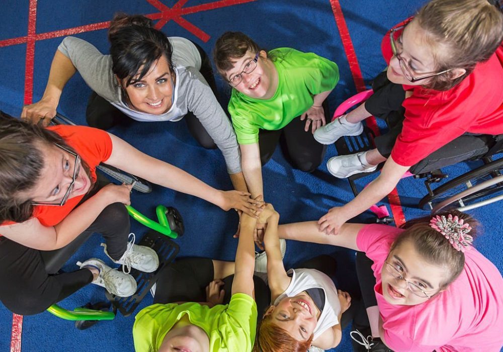 Overhead view of a group of special needs teenagers and young women with instructor, showing team spirit. They are sitting in a circle on a gym floor, hands in the center. Several of the girls have downs syndrome and two are in wheelchairs.